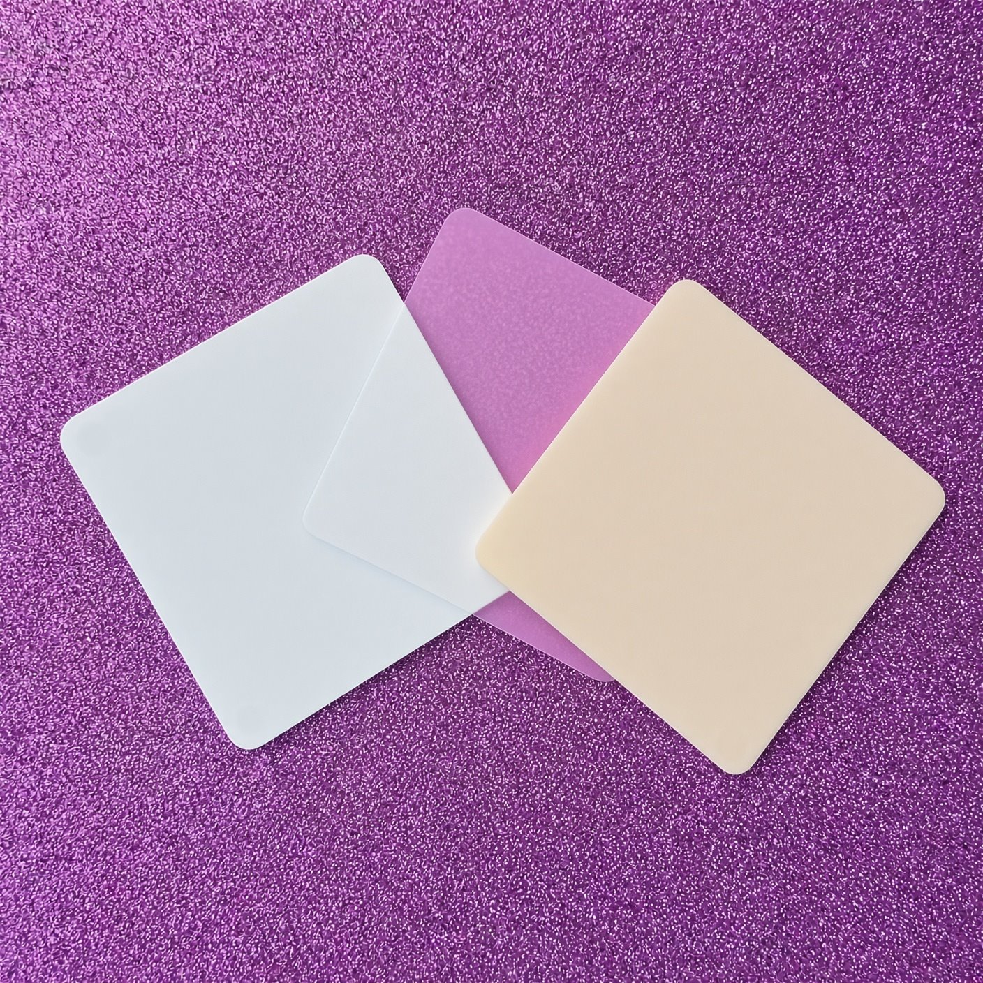 Drei Zuschnitte aus PLEXIGLAS® auf lila Glitzeruntergrund: links Weiß, in der Mitte milchig, rechts Creme, durch die milchige Platte schimmert der Untergrund