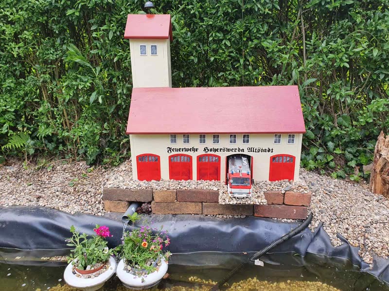 Feuerwehrhaus-Modell frontal am Gartenteich, ein Löschfahrzeug fährt aus dem Tor