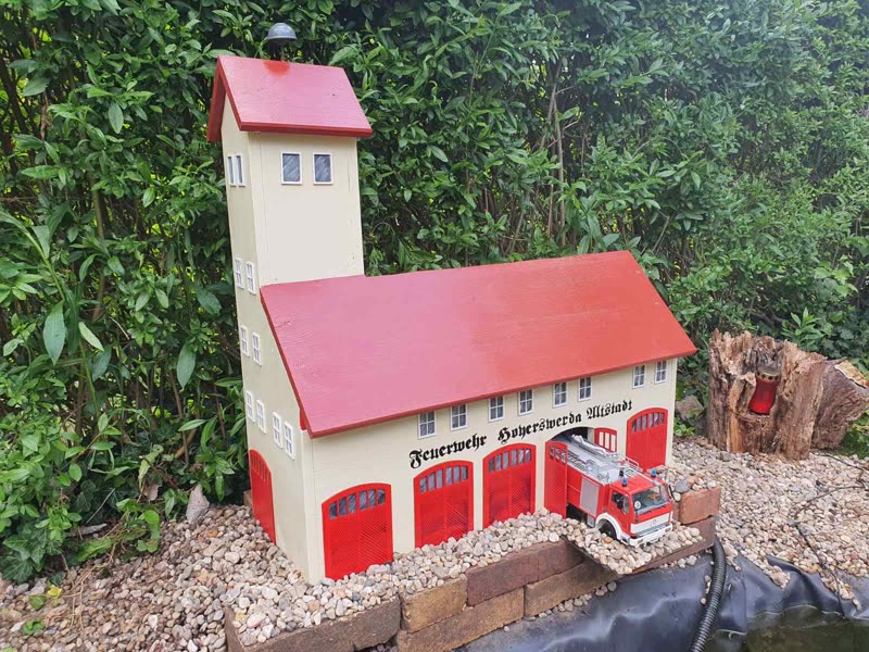 Modell eines Feuerwehrgerätehauses mit Schlauchturm, rotem Dach und roten Toren im Garten